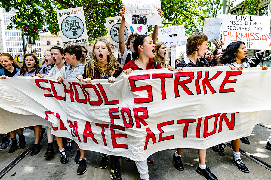 Photograph shows young people marching in the street with a banner that says “School strike for climate action.” They are holding various placards.