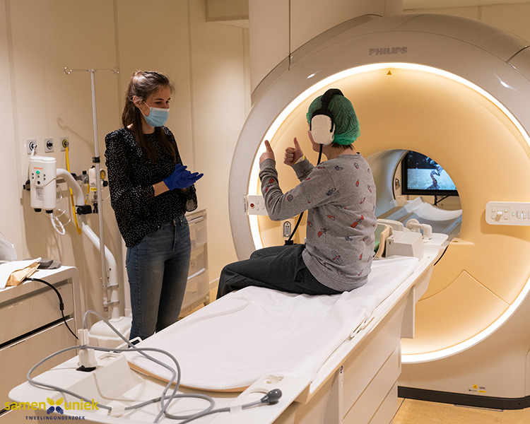 Photo of an adolescent sitting on the table of an MRI scanner holding their hands up; the child wears a long-sleeved shirt, hairnet and earphones and is talking to an adult standing nearby. Both wear masks. An MRI scanner is in the background.