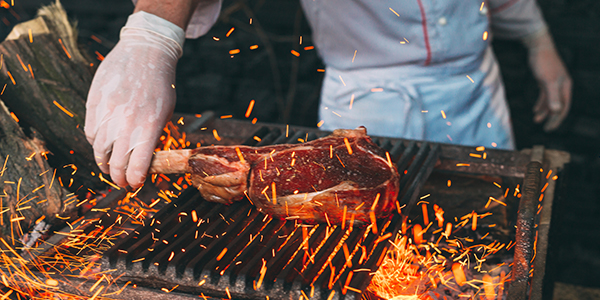 A cook grills a large, thick-cut beef steak on a charcoal fire.