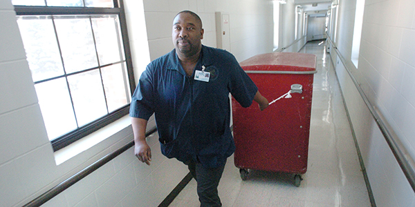 Photo shows a Black hospital worker pulling a trash container down a hallway.