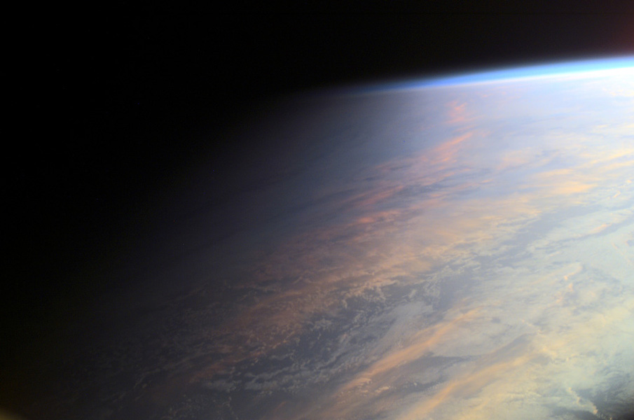 Photo shows a portion of Earth bathed in twilight. The sun illuminates clouds and ocean on the right through diffuse, hazy light that transitions to darkness on the left.