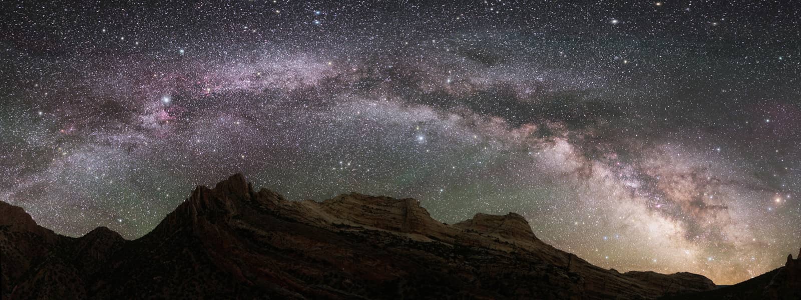 Photograph at night, with mountainous terrain and desert floor. In the sky one can see thousands of stars and the disk of the Milky Way galaxy.