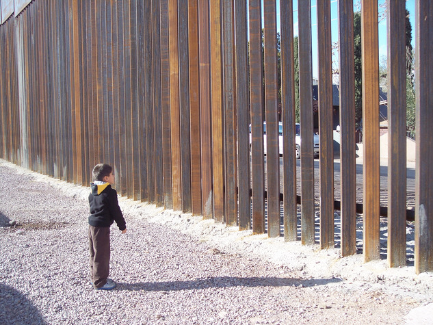 Photo of a small boy facing a border wall made of vertical slats, with spaces between.
