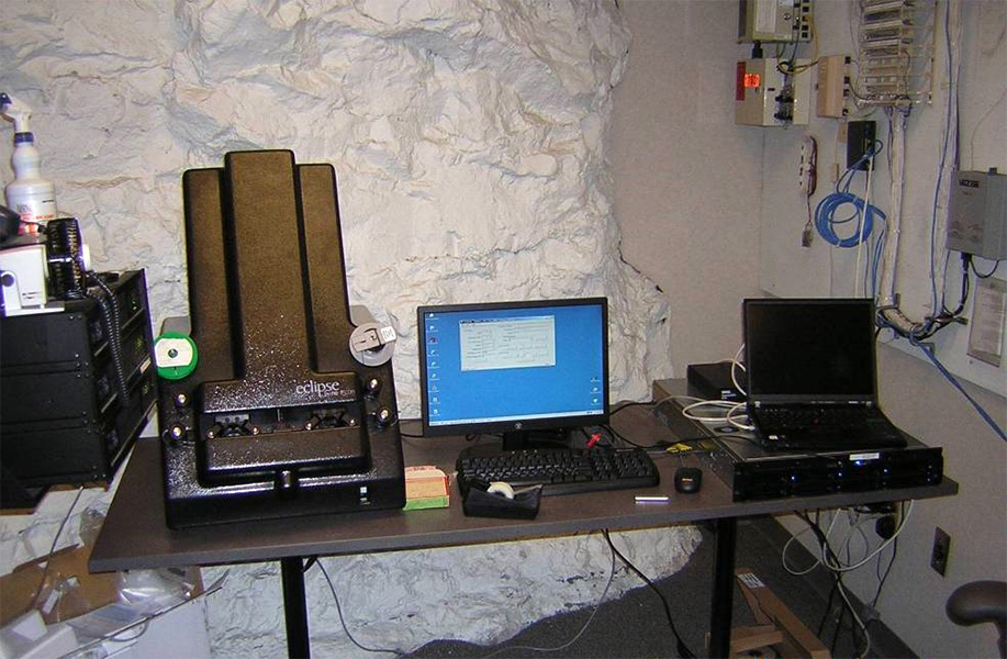 Photo of a desk with a computer and other office items in front of an uneven rock wall that is painted white.