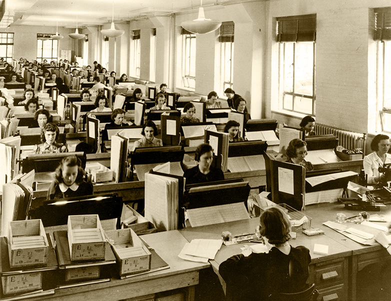 Black-and-white photo of women sitting at rows of desks entering data, surrounded by documents and books.