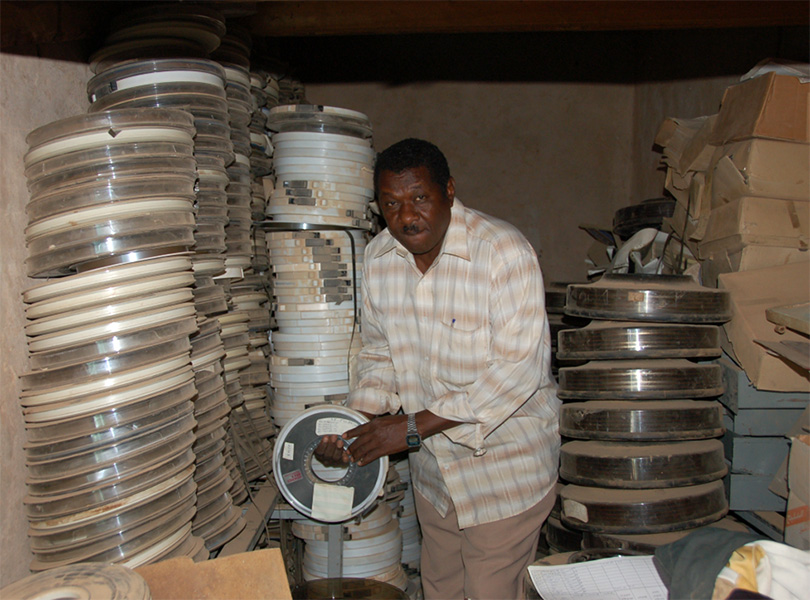 Photo of a man in low-ceiling room surrounded by tall stacks of dusty film reels, one of which he holds in his hand.