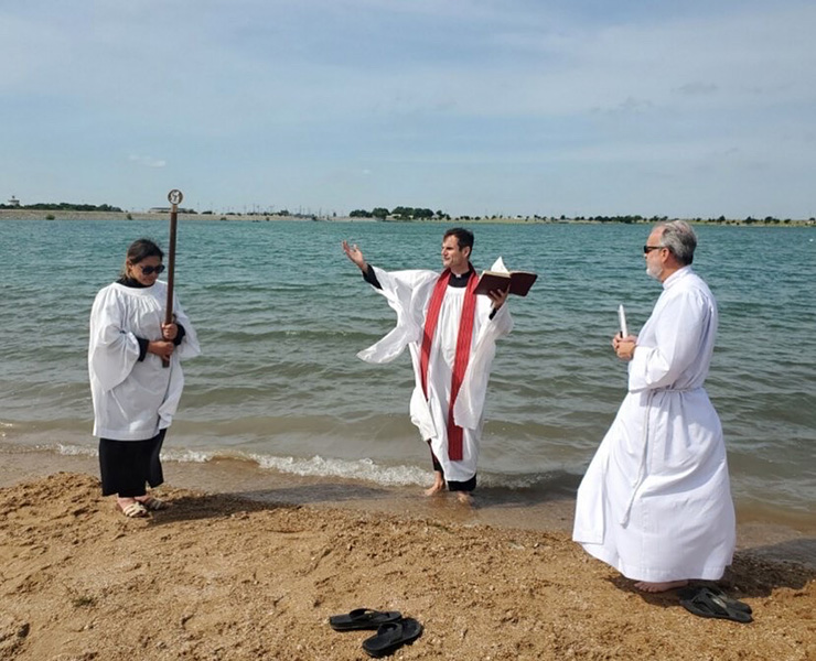 Photograph of a man wearing a white minister’s robe and a red stole, standing by water. One hand holds a book and the other hand is raised in the air. To each side of him stand two other people in white robes holding religious items.