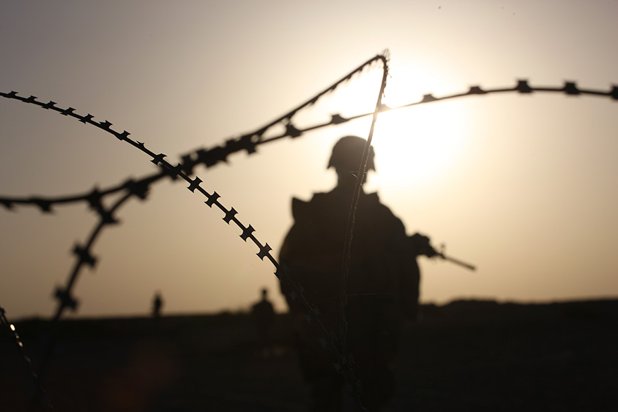 Photograph shows silhouette, from behind, of a helmeted soldier holding a gun. Barbed wire is to the front.
