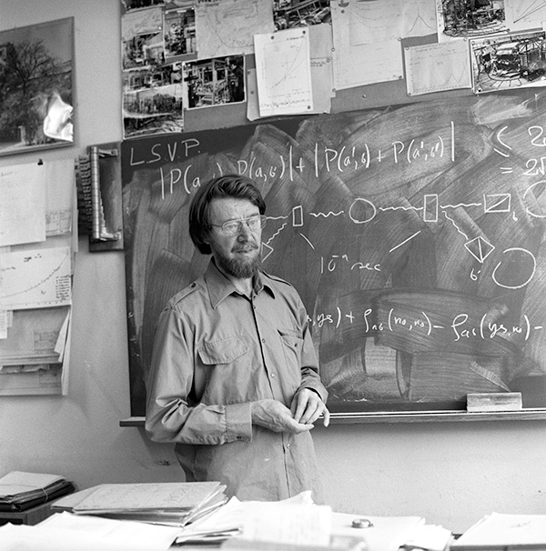 Historic photo shows John Bell standing in front of a chalkboard with equations and drawings on it, with papers piled on a table in front of him and tacked to the wall around the chalkboard.