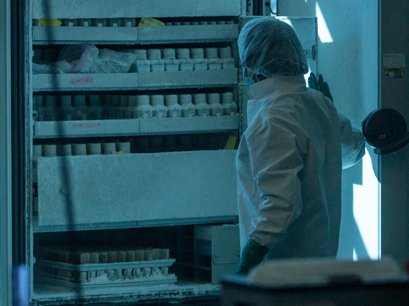 Photograph of a technician standing next to a freezer with its door open. Shelves of containers holding stool samples are stacked inside the freezer.