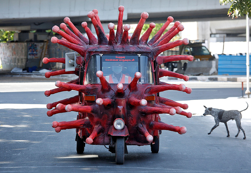 Photograph of an autorickshaw covered with coronavirus-like spikes. A man is driving it down a street; a dog looks on.
