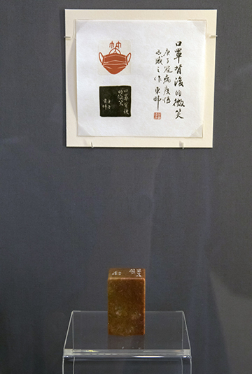 Photograph of a museum exhibit. A carved seal is standing on a small glass table. Above it is a printed image with Chinese calligraphy.