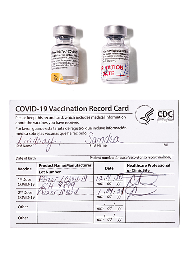 A photograph shows two small vaccine vials and the vaccine card for Sandra Lindsay, filled out with the dates of her two vaccine doses.