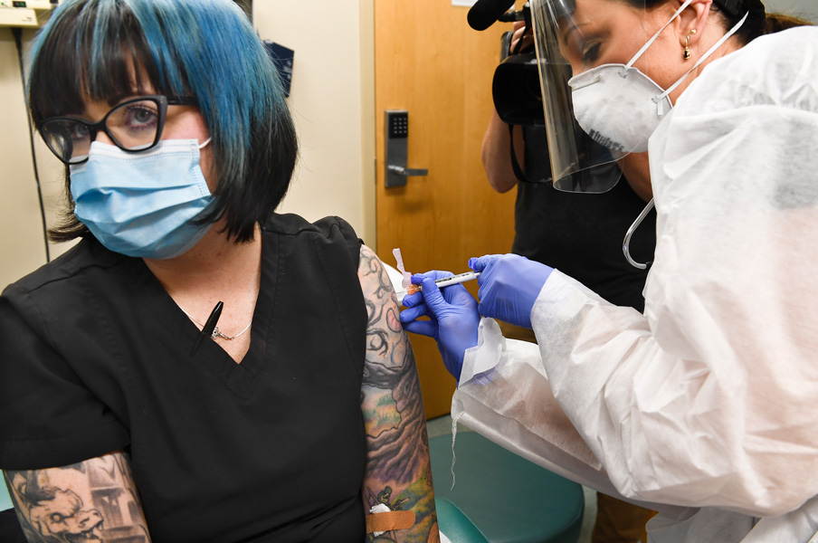 Photograph of a woman wearing a black, short-sleeve top and blue surgical mask receiving an injection in her left arm by a nurse wearing blue gloves, N95 mask, face shield and white gown.