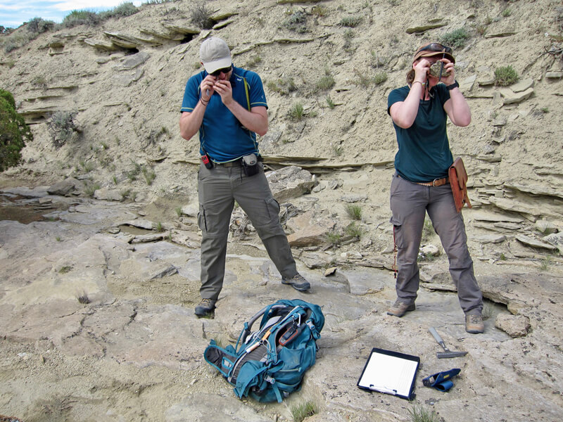 Photograph of Joe Bonsor on the left and Susie Maidment on the right. They are standing on rock, with a rock face behind them, holding hand lenses close to their faces. A backpack, logbook and tools are at their feet.