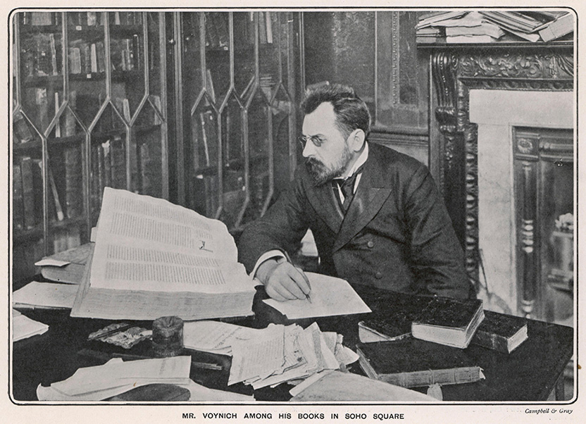 Old photograph of a man with a beard sitting at a desk covered with books and papers. He is writing something on a piece of paper and looking intently at a large book.