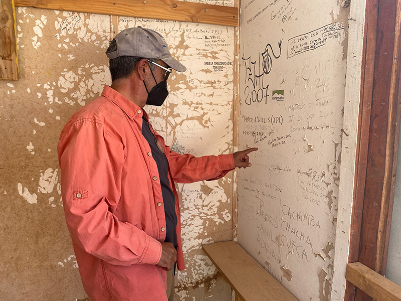 A man points to a wall with peeling paint; people’s names are scribbled all over the wall