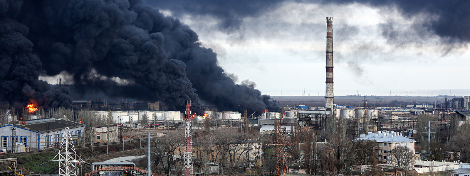 A photograph shows huge clouds of black smoke darkening the sky above the containers and other structures of an oil storage facility. Flames are also visible.