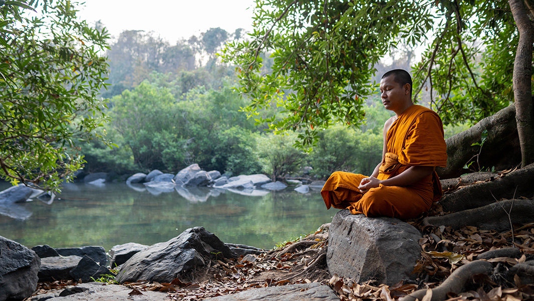 A man clad in orange is sitting on a rock with his eyes closed, meditating. In the background are a river, rocks and trees.