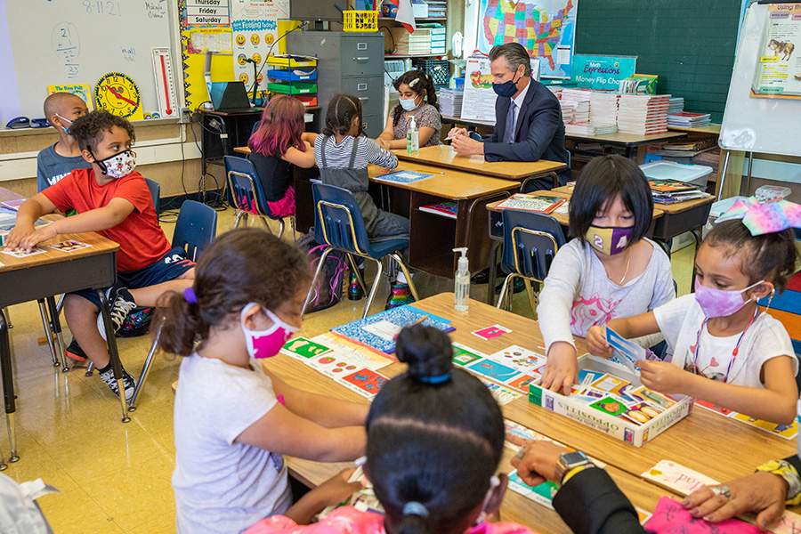 Photograph of an elementary school classroom. Children wearing colorful masks are sitting at tables. Governor Gavin Newsom, wearing a suit and dark mask, is sitting at one of the tables.