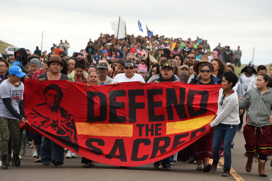 Photograph of a group of demonstrators. In the front, people carry a large banner that declares “Defend the sacred.”