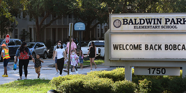 Photograph of parents and children walking toward a school. Some are wearing masks, some art not. Kids are carrying backpacks. A school crossing attendant wearing an orange vest and holding a stop si