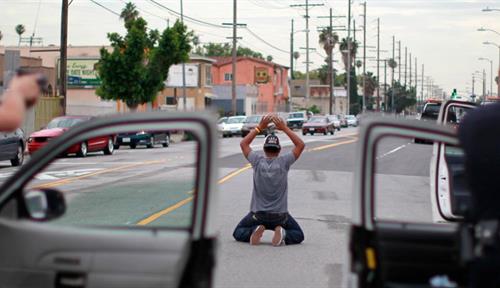 Photograph of a man kneeling on the ground on a city street, with his hands above his head. Two police officers stand by their cars, guns drawn.