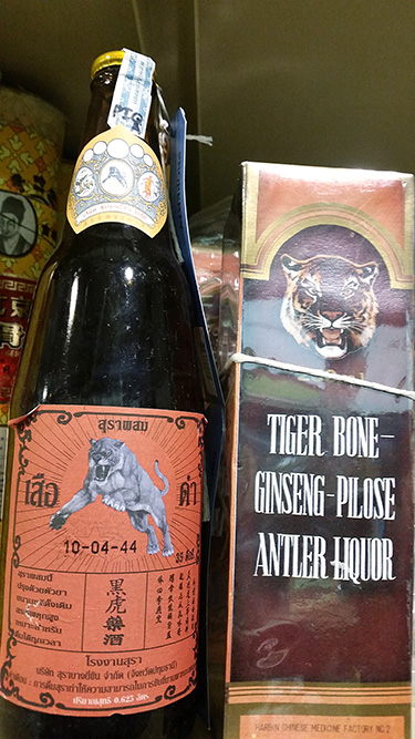 A photograph shows two products. On the left is a bottle with a label showing a leaping big cat. On the right is a box with a picture of a tiger face and the label “TIGER BONE-GINSENG-PILoSE ANTLER LIQUOR.”