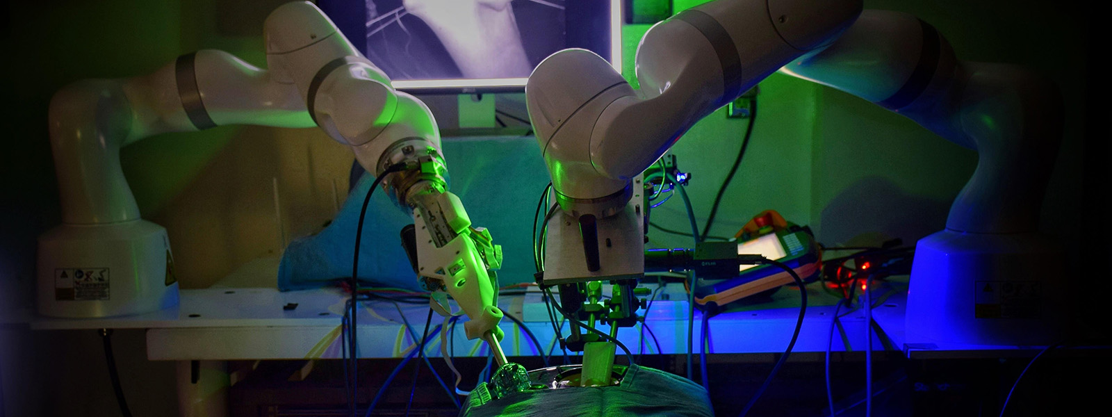Two robotic arms hover over a draped form. In background a monitor shows a length of intestine gripped by clamps.