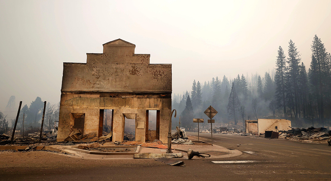 A burned-out storefront is all that remains standing at a street corner ravaged by wildfire. Smoke hangs in the background.