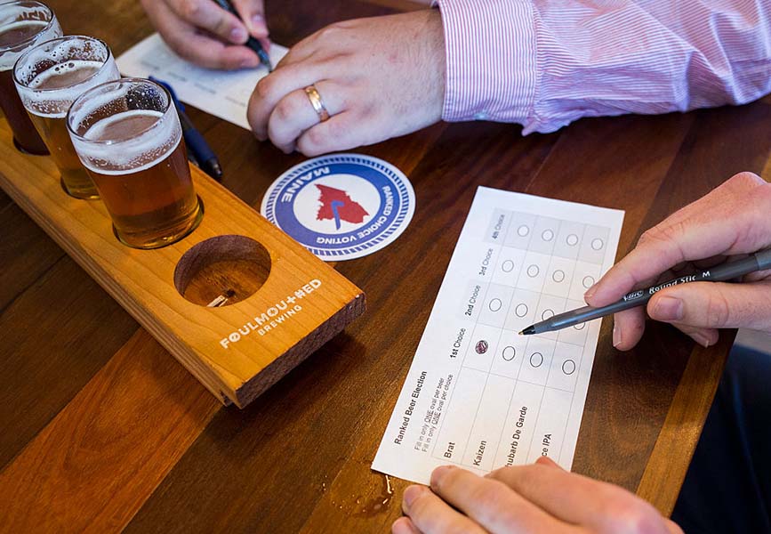 Photograph of several glasses of beer in a beer flight holder. Pairs of hands of two people are shown close up as they fill out ballot cards to rank the beers.