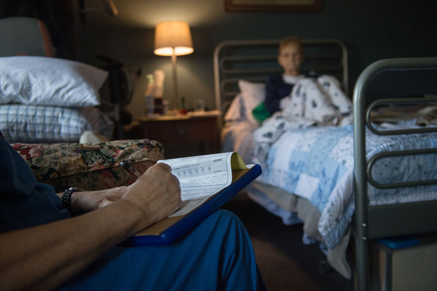 A woman lies in a hospice bed talking to a medical worker, who makes notes on a piece of paper.