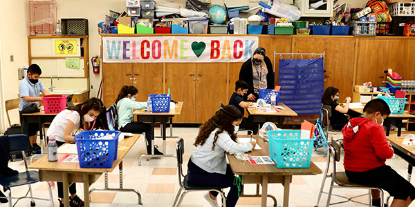 Elementary school students wear face masks while they sit at their own desks in a classroom in Los Angeles in April 2021. Their masked teacher walks amongst them, with a Welcome Back sign on the wall 