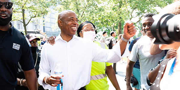 Photograph of Eric Adams smiling and raising his fist in victory, surrounded by other people, some of whom are also smiling.