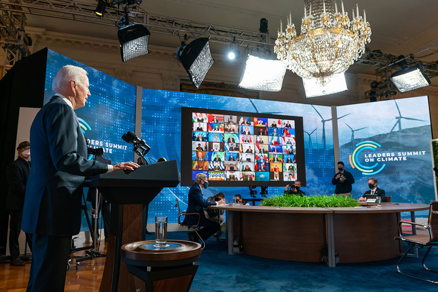 Photo shows President Joe Biden at a podium speaking to an offscreen camera at the Leaders Summit on Climate Session 1: “Raising Our Climate Ambition” on April 22, 2021. John Kerry and other White House staff sit at a nearby conference table and dozens of world leaders participate via video chat on a large monitor.