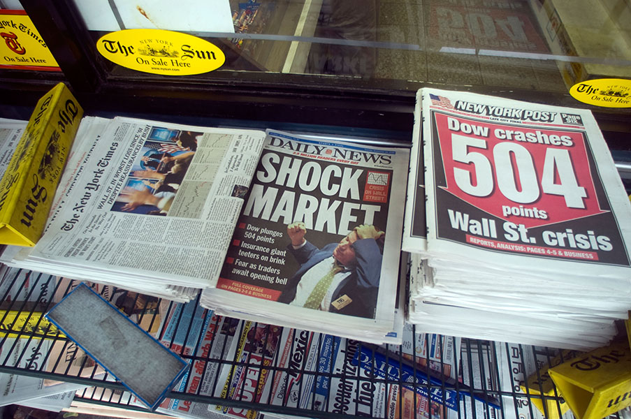 Photo shows bundles of newspapers at a news stand on September 16, 2008, with large headlines about the falling stock market: “Shock Market” (Daily News) and “Dow Crashes 504 Points: Wall St. Crisis” (New York Post).