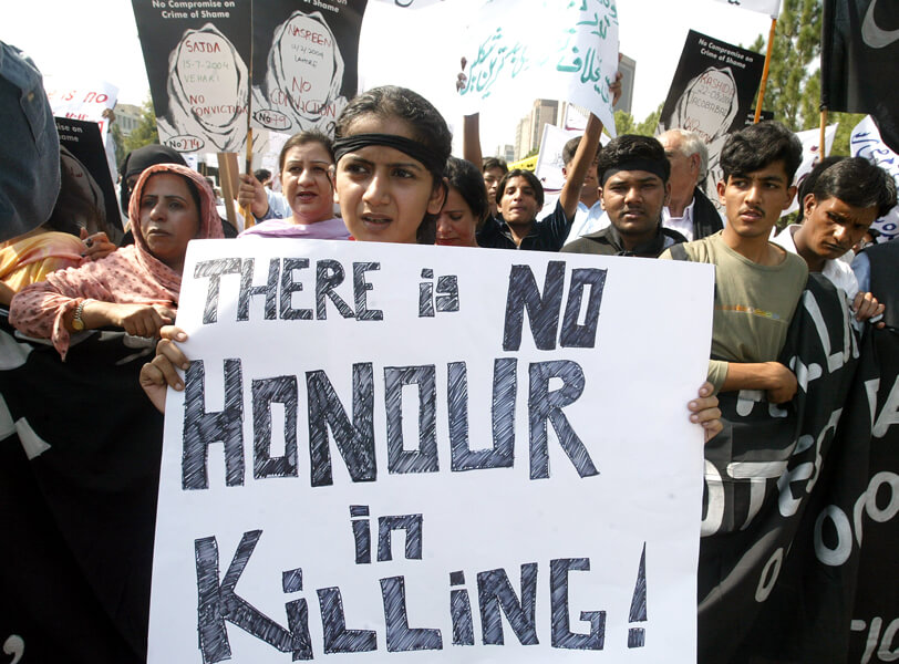 Photograph of a demonstration. Women and men hold placards in the air. At the front, a woman holds a large sign that says, “There is no honour in killing!”