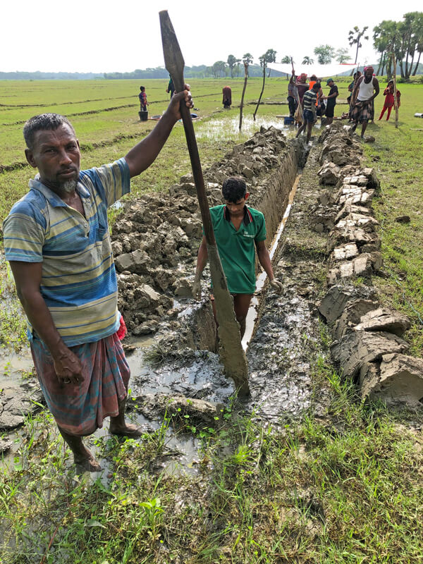 Photo shows people digging a trench for a GPS cable, in a green field. In the foreground, a man in the trench holds a long wooden tool and looks at the camera.