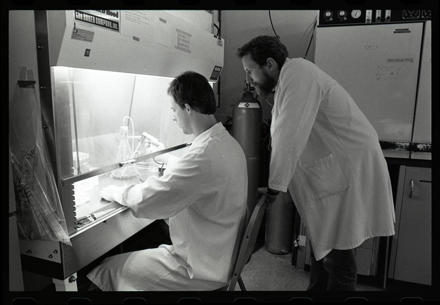 Black and white photo of two men in front of a lab hood. One is sitting, working in the hood. Charles Rice is standing, watching, to the right.