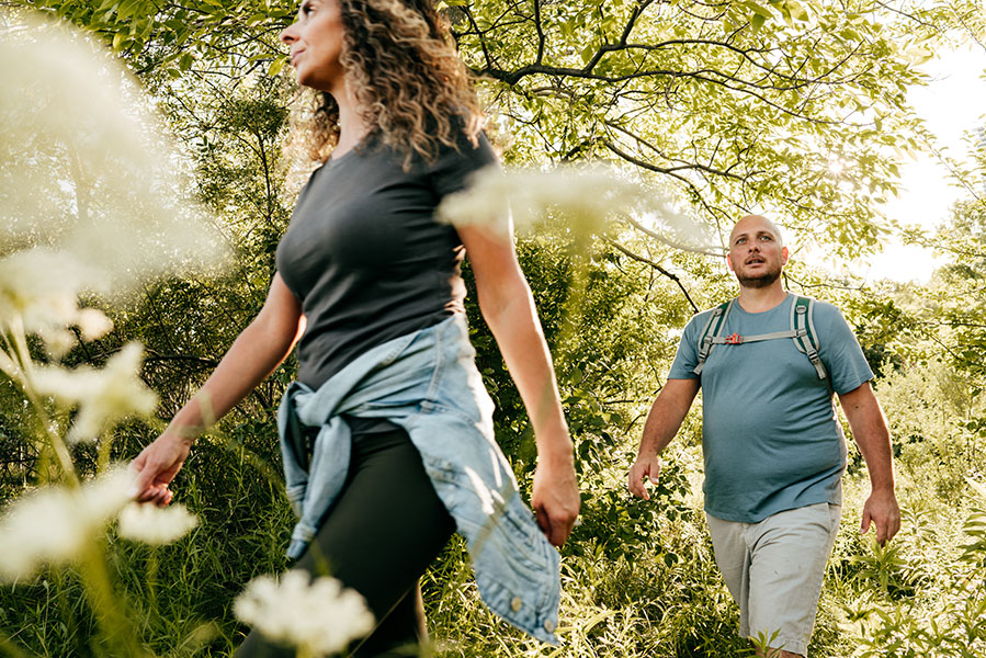 A fit-looking woman and an overweight man walk through the woods.