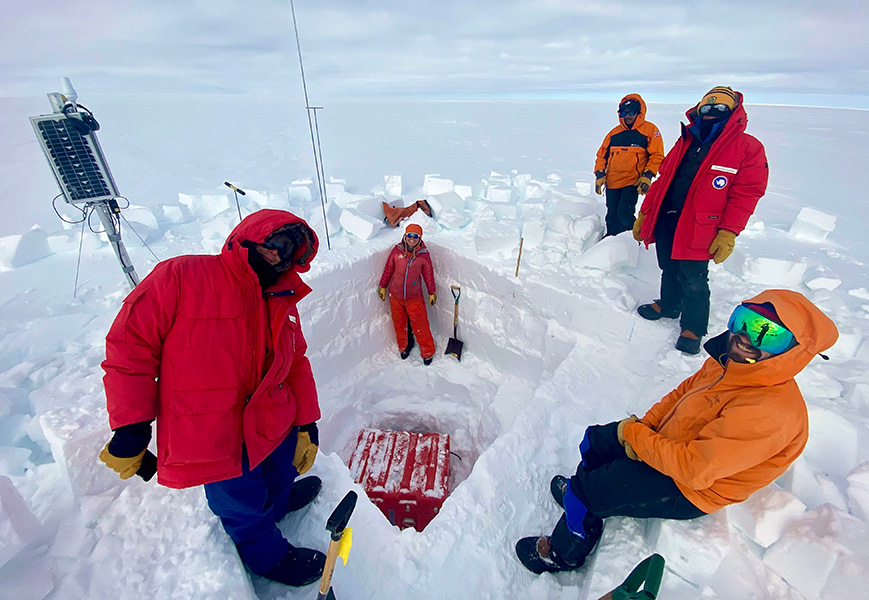 Several people in cold-weather clothing pose near a hole they’ve dug in a glacier, with various pieces of equipment nearby.