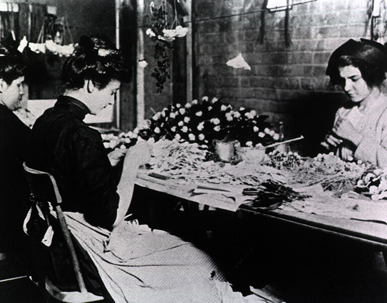 Black-and-white photo of three women sitting at a table making flowers for the garment industry.