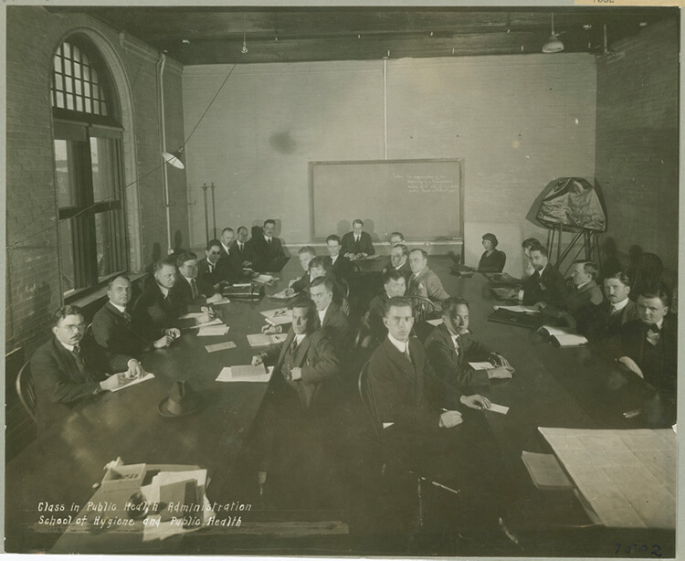 Black-and-white photo of several men seated around two tables in front of a blackboard for a class in public health administration.