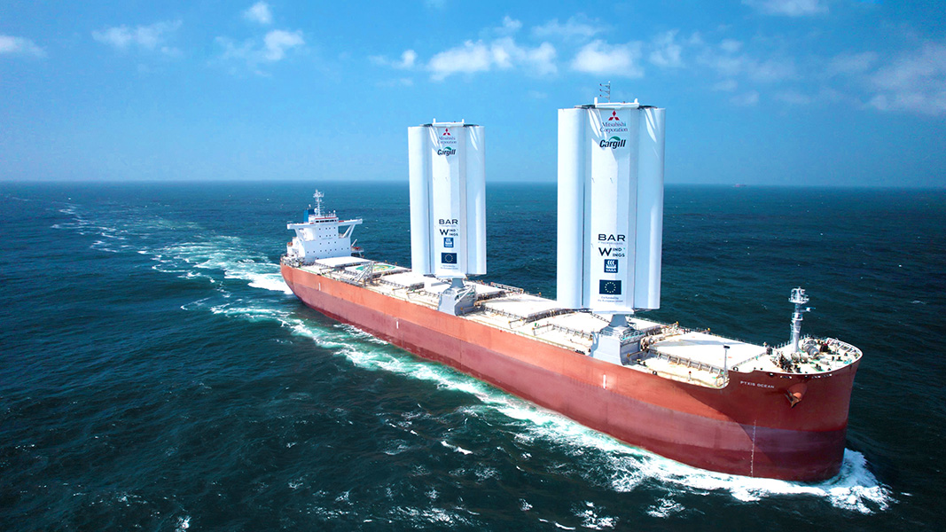 Photograph of a ship in the water, with rust-red hull and two large structures — the sails — that look like several upright cylinders joined together.