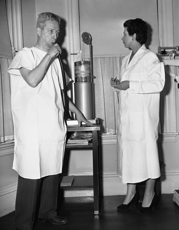 Black and white photograph of a man in a gown blowing into a tube as a technician in a lab coat stands watching on the right.