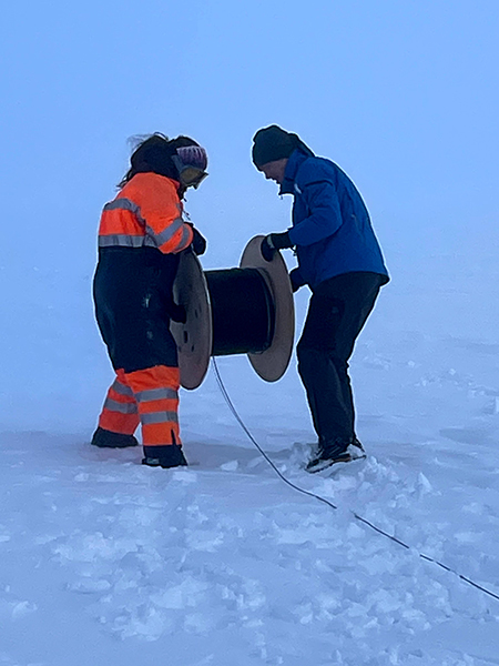 Photograph of two warmly dressed workers holding a large spool and unrolling cable into the snow.