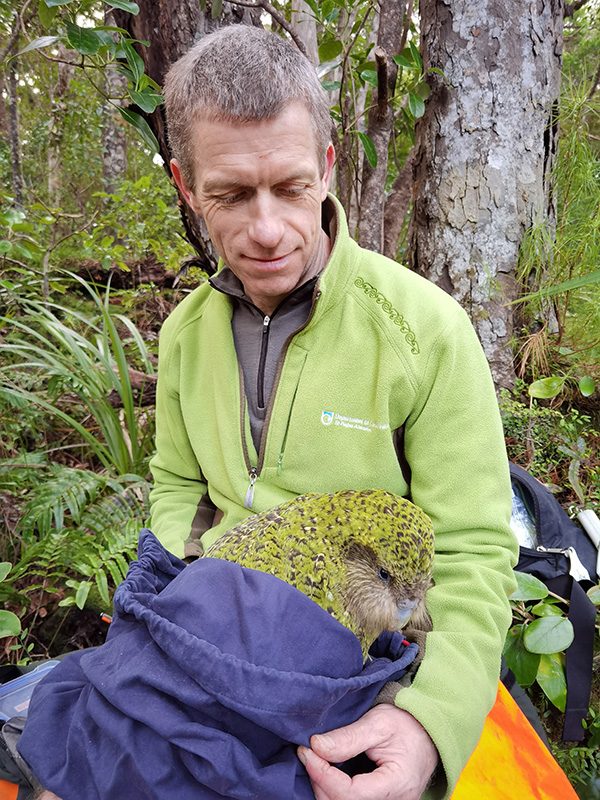 Conservation biologist Andrew Digby sits in a forest holding a kākāpō swaddled in blue cloth.