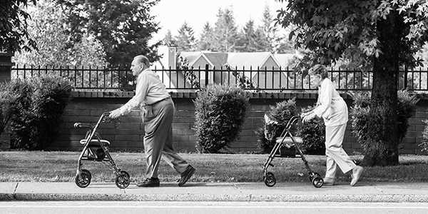 Photo of an old man and woman, each pushing a wheeled walker, strolling down a sidewalk.