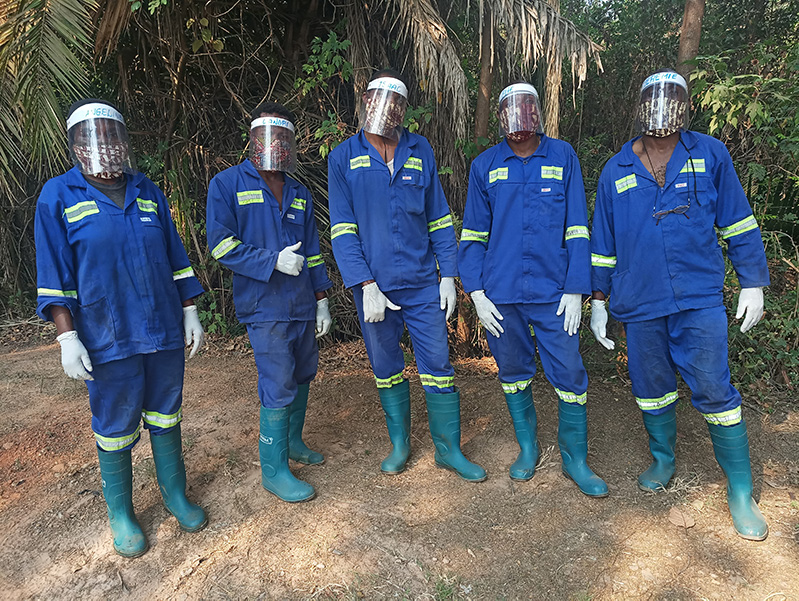 Photograph shows five people standing in front of some vegetation and palm trees. They are wearing blue work uniforms, face shields and tall rubber boots.
