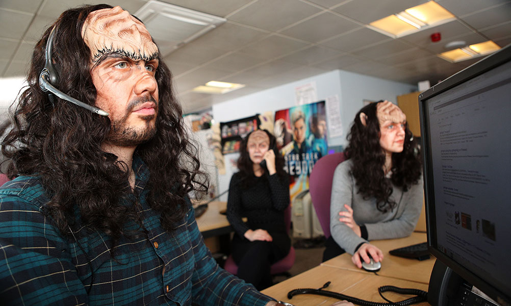 Several people with their faces made up to look like Klingons sit at computers and talk on headsets.